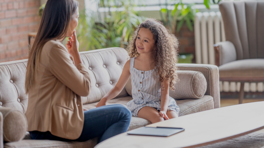Una mujer ofrece terapia de lenguaje para niños a una niña pequeña con cabello rizado, sentadas juntas en un sofá beige en una oficina luminosa con plantas y una pared de ladrillo, representando servicios en Monterrey.